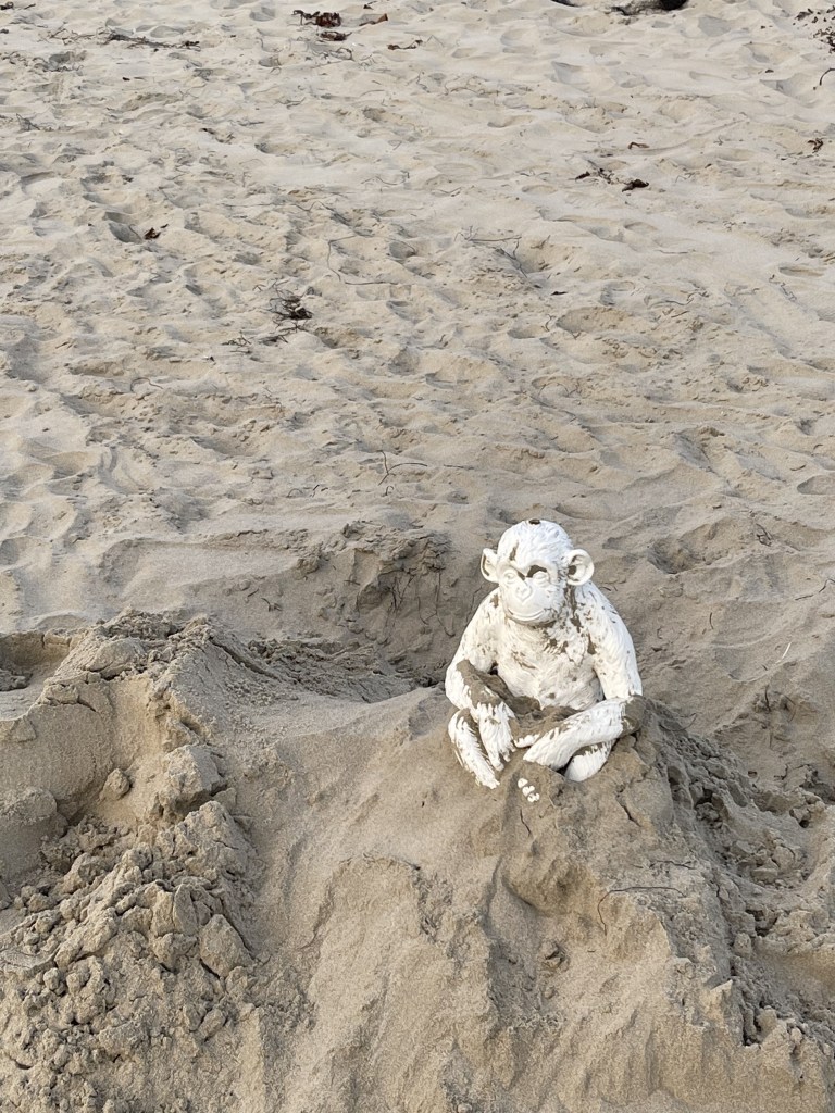 White plaster monkey statue sits on a hill of sand. He smiles as he takes in the view.